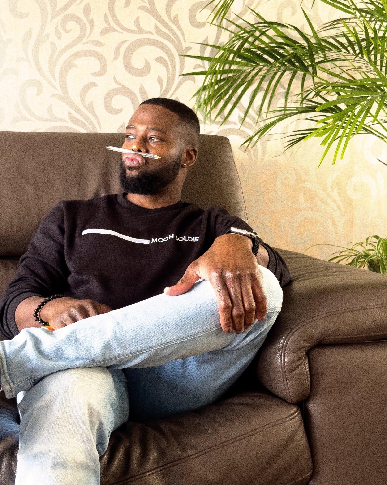 Black soft cotton sweatshirt featuring Moon Soldier's minimalist white wave design on the chest, worn by a man seated on a brown leather couch with a casual pose, surrounded by indoor greenery.