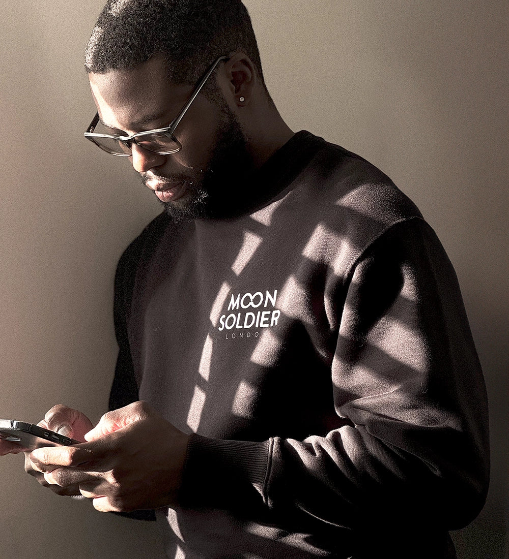 Man wearing a Moon Soldier black soft cotton crewneck sweatshirt, standing in soft lighting while holding a phone.