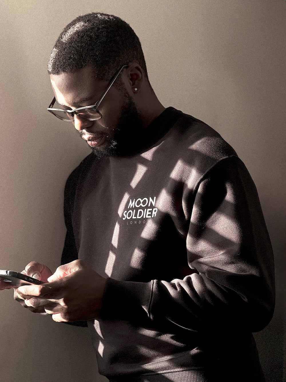 Man wearing a Moon Soldier black soft cotton crewneck sweatshirt, standing in soft lighting while holding a phone.