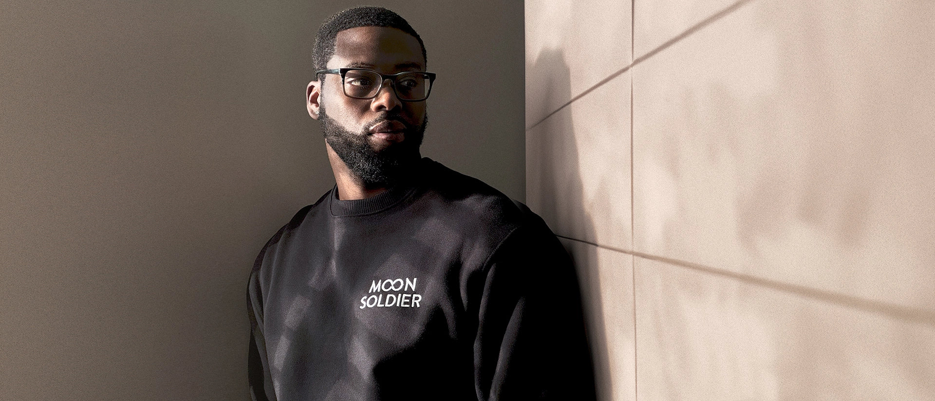 Model wearing a black soft cotton heavy sweatshirt with the Moon Soldier logo, photographed indoors with warm lighting and subtle shadow effects on the wall.