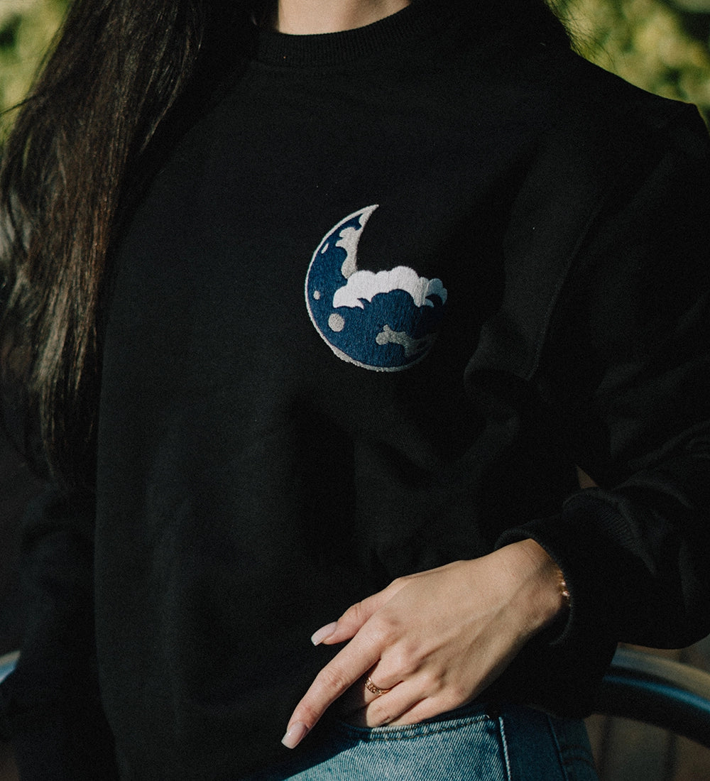 Black soft cotton sweatshirt featuring an embroidered blue moon logo on the chest, modeled outdoors with a casual denim pairing.