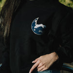 Black soft cotton sweatshirt featuring an embroidered blue moon logo on the chest, modeled outdoors with a casual denim pairing.