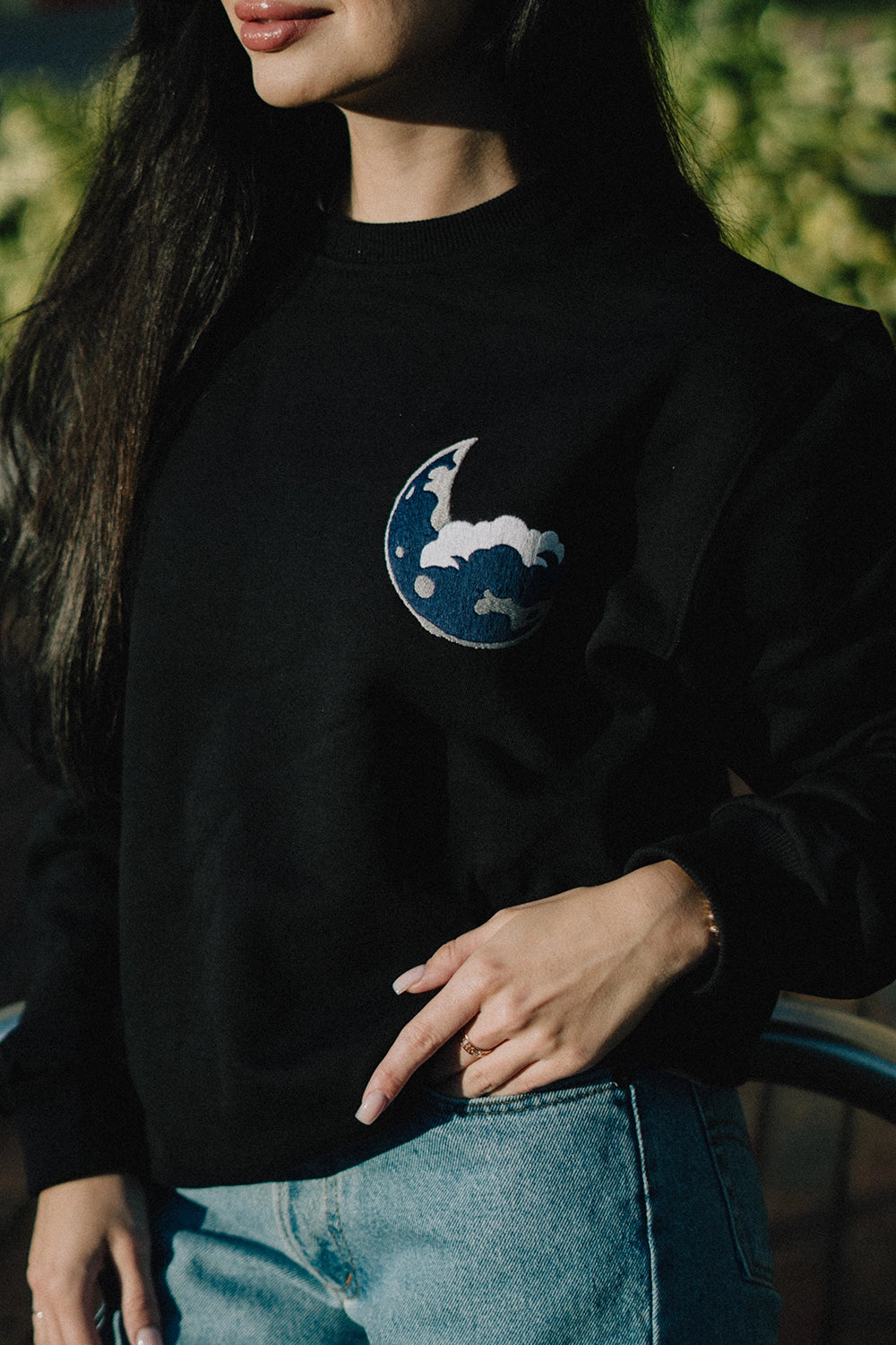 Black soft cotton sweatshirt featuring an embroidered blue moon logo on the chest, modeled outdoors with a casual denim pairing.