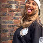 Smiling model wearing a black Moon Soldier soft cotton sweatshirt with a melted moon design and QR code badge, styled with a white beanie in an outdoor urban setting.