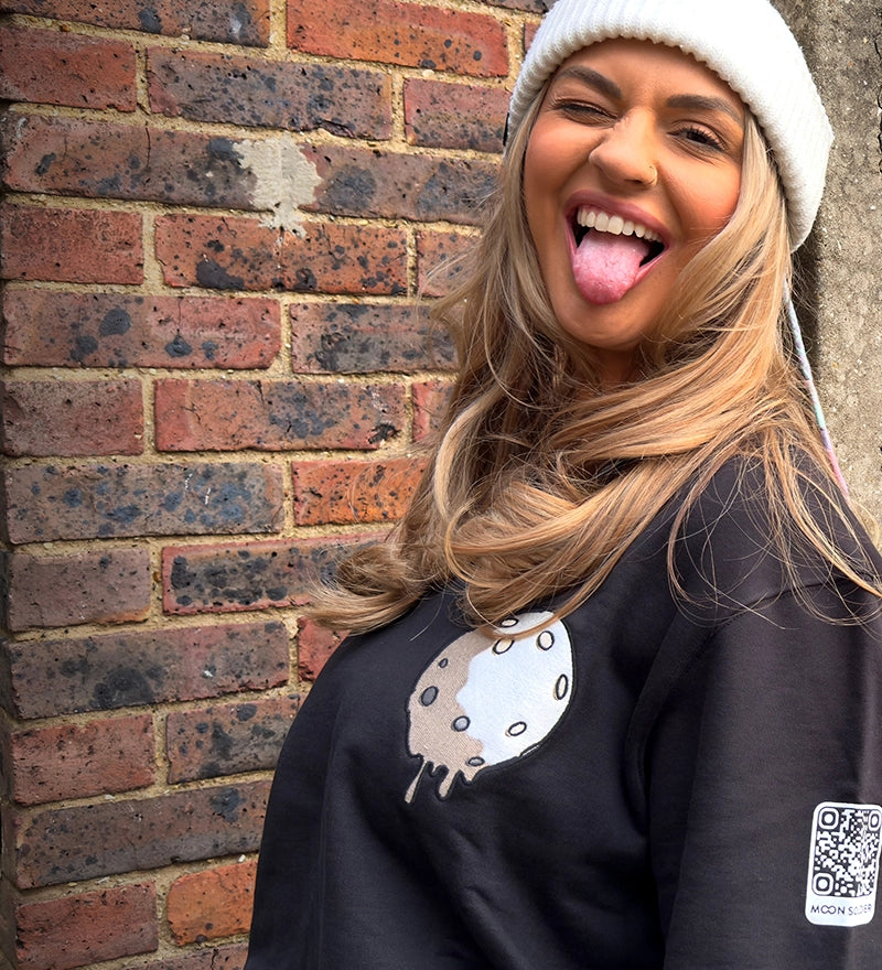 Smiling model wearing a black Moon Soldier soft cotton sweatshirt with a melted moon design and QR code badge, styled with a white beanie in an outdoor urban setting.