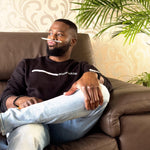 Black soft cotton sweatshirt featuring Moon Soldier's minimalist white wave design on the chest, worn by a man seated on a brown leather couch with a casual pose, surrounded by indoor greenery.