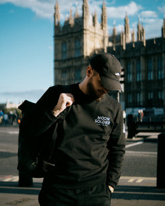 Model wearing Moon Soldier black sweatshirt with logo in front of historic building, carrying a backpack in an urban setting.